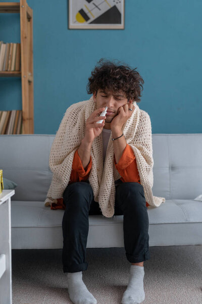Sick young curly man spraying his nose with nasal spray sitting on sofa at home. People and health problem concept