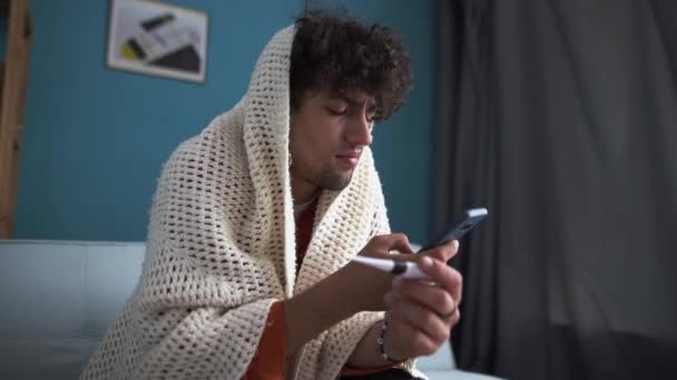 毛布に覆われた不健康なアラビア人は スマートフォンの医師と高温の話をオンラインで相談測定します 病気の男はインフルエンザに苦しんでいます 医療の概念 — ストック動画