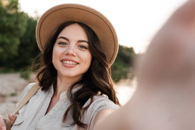 Genç bir kadın dışarıda selfie portresi çekiyor. Kumsalda yaz tatilinin tadını çıkaran şapkalı mutlu bir kız. Yakın plan.
