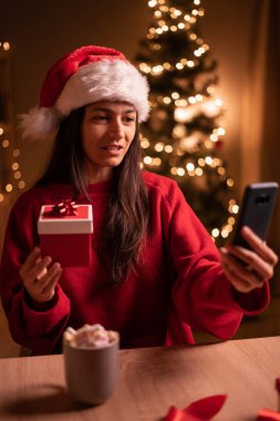 Mutlu İspanyol kadın Noel arifesinde evde akıllı telefondan video çekerken hediye kutusu gösteriyor. Boşluğu kopyala