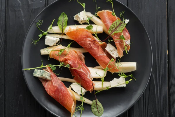 stock image Appetizer of prosciutto, pear and blue cheese on a black plate for the holiday, close-up, top view. Copy space