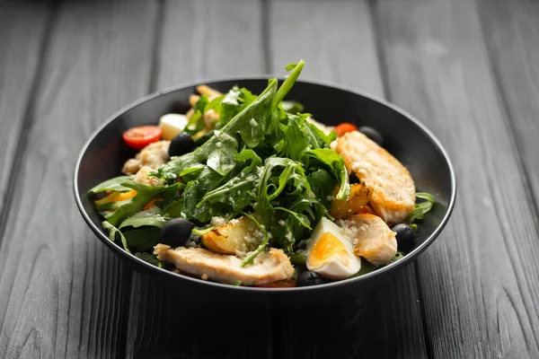 stock image Caesar salad with fried chicken breast, egg, arugula leaves, cherry tomatoes and olives on a dark wooden background. Copy space