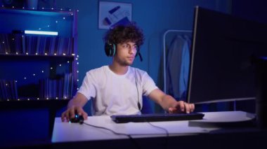 Joyful Arabic gamer rejoices in winning a video game, wears headset sitting at home at night with neon light. Streamer winner plays at home on a personal computer. Copy space