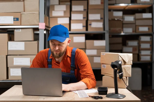 Serious male worker sitting and working with computer laptop in storage warehouse. People, warehouse and industry concept. Copy space