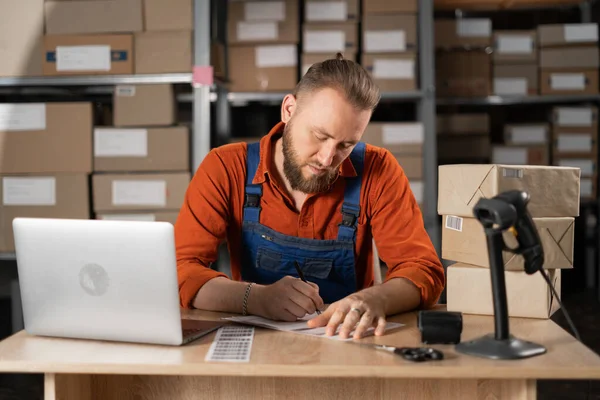 Inventory manager small business owner working on laptop in warehouse, writing delivery address on sticker.Boxes background. Copy space