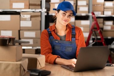 Depoda oturmuş teslimat için paket hazırlarken dizüstü bilgisayarda çalışan mutlu bir işletme sahibinin portresi. Bir kadın posta deposunda gülümsüyor ve kameraya bakıyor. Boşluğu kopyala