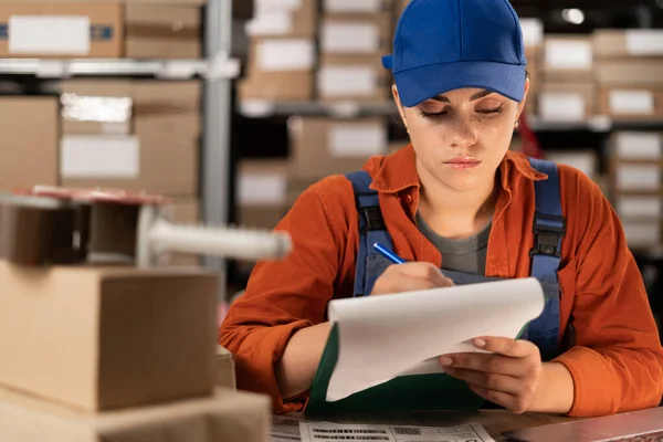 Portrait of worker in warehouse preparing goods for dispatch writing information in clipboard. Copy space