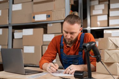 Profesyonel bir erkek işçi dizüstü bilgisayarını kullanarak malzeme dolu raflarla dolu depoda stokları ve envanterleri kontrol ediyor, teslimat için adres yazıyor. Boşluğu kopyala