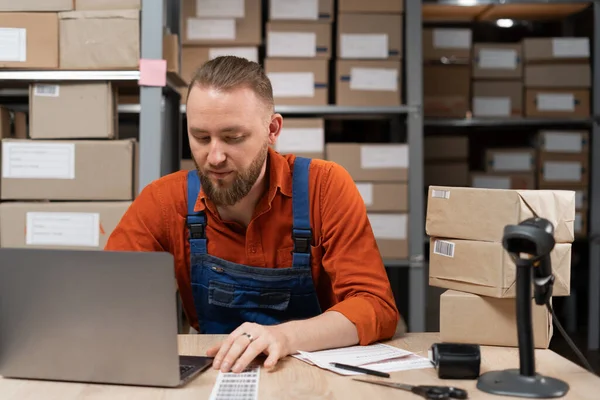 Manager using laptop computer to check inventory while working in warehouse retail center with cardboard boxes. E-Commerce concept