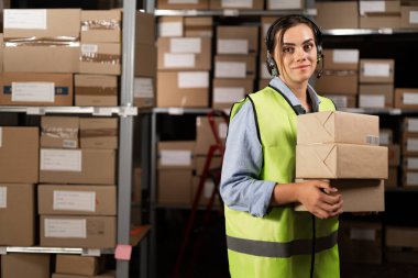 Telsiz kulaklıklı, yansıtıcı yelek giymiş genç bir kadın lojistik deposunda elinde karton kutularla kameraya bakıyor. Boşluğu kopyala.