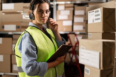 Lojistik deposunda dijital tablet kullanan Brezilyalı kadın çalışan. Arkaplanda karton kutular olan raflar kameraya bakıyor. Boşluğu kopyala.