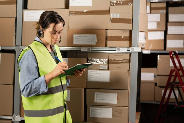 A professional female worker in a reflective vest checks inventory and inventory with a clipboard in a retail warehouse full of product shelves. People working in logistics distribution center