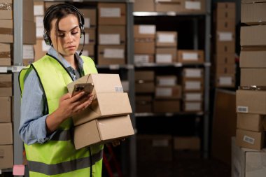 Telsiz kulaklıklı, yansıtıcı yelek giymiş bir kadın lojistik deposunda elinde karton kutularla kameraya bakıyor. Haberlere göz atıyorum. Boşluğu kopyala.