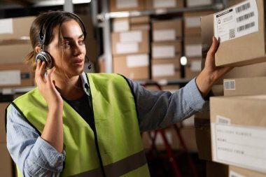 Bir lojistik deposunda dijital tablet kullanarak gerekli ürünü arayan Brezilyalı bir kadın işçi. karton kutularla rafların arka planına karşı.