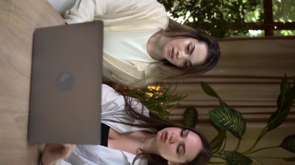 リモートチーム2人の女性が カフェでラップトップを使用して一緒にビジネスプロジェクトを議論し 作業しています 若いビジネスマンは 彼らのコラボレーションのためのアイデアをブレインストーミングします バーティカルビデオ — ストック動画