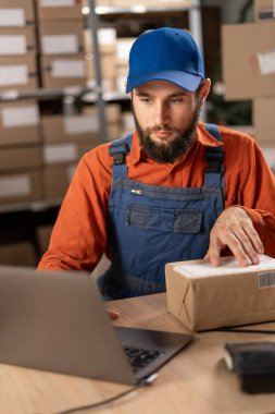 Depo yöneticisi dizüstü bilgisayarı kullanarak bir paketin sipariş numarasını kontrol ediyor, karton kutuyu posta ücreti için hazırlıyor. Boşluğu kopyala