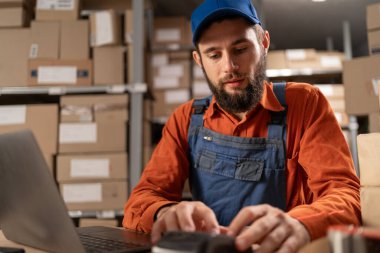 Depoda karton kutularla, online e-ticaret siparişleriyle çalışan genç adam ödeme için dizüstü bilgisayar ve pos-terminal kullanıyor. Yakın plan.