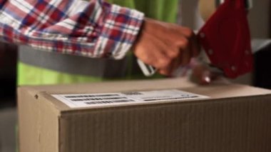 Perakende satış mağazasındaki bir depoda yelek giyen işçiler yapışkan bantla bir kutu paketlemişler. Boşluğu kopyala