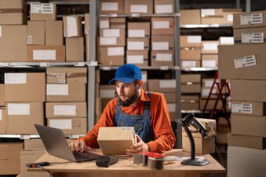 Bir paketin sipariş numarasını kontrol etmek için envanter erkek çalışanı dizüstü bilgisayar kullanıyor, posta ve teslimat için karton kutu hazırlıyor. Depoda çalışma günü. Boşluğu kopyala
