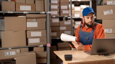 Depo müdürü büyük bir depoda dizüstü bilgisayar ve pano kullanarak paket listesini kontrol ediyor. Boşluğu kopyala