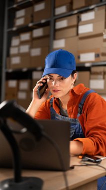 Depoda dizüstü bilgisayarla çalışan bir kadın küçük işletme sahibi telefonda konuşuyor. Girişimci elinde cep telefonu ile depoda perakende satış paketi hazırlıyor. Dikey