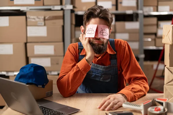 Exhausted tired warehouse worker with painted eyes on stickers, adhesive notes on face sleeping at workplace, sitting at desk with laptop, lazy young male working on difficult project. Copy space