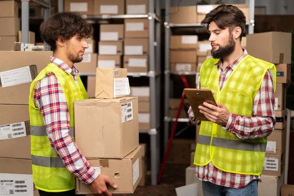Male inventory manager shows digital tablet information to a arabic worker holding cardboard boxes, talking about work. Stock of Parcels with Products Ready for Shipment on background. Copy space