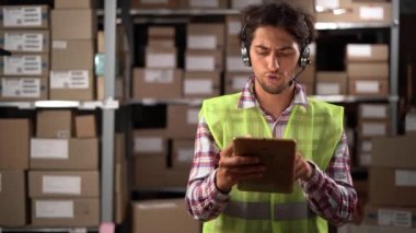 Lojistik dağıtım deposunda envanter veya kontrol için dijital tablet kullanan kulaklık takan bir erkek işçi. Boşluğu kopyala