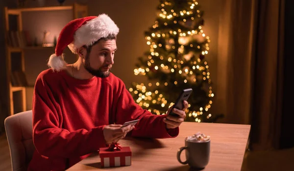 サンタの若者は クリスマスプレゼントのクレジットカードでオンラインで支払い スマートフォンを使用して オンラインショッピング ホームオフィスで冬の休暇を楽しんでいます コピースペース — ストック写真