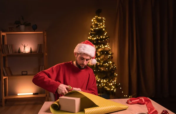 テーブルに座ってクリスマスプレゼントを準備する若者は 夕方に自宅のオフィスでハサミで包装紙を切り取ります コピースペース — ストック写真