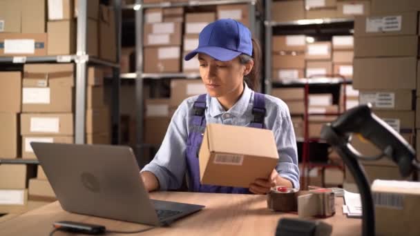 ラップトップコンピュータを使用して小包の注文番号を確認し 郵便のための箱を準備する在庫の女性の労働者 背景に箱のパッケージが付いている棚 コピースペース — ストック動画