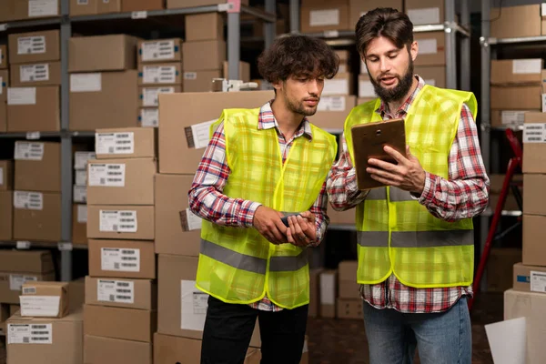 Hispanic male inventory manager shows tablet information to a worker talking about work. Stock of parcels with products for shipment on background. Copy space