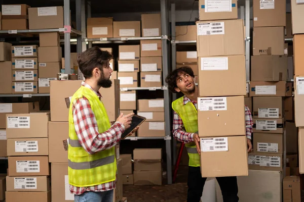Two warehouse mixed race workers using a digital tablet while recording inventory and preparing shipment. Logistics employees working with warehouse management software in a large distribution center.