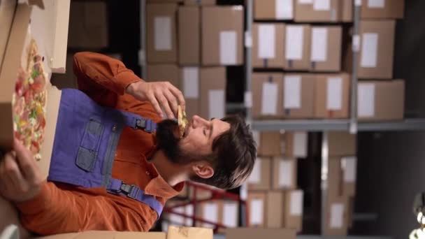 男性の倉庫労働者は 休憩で楽しむランチを楽しんでピザを食べています バーティカルビデオ — ストック動画