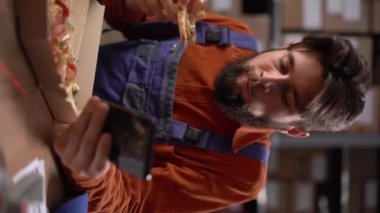Depo işçisi oturur ve dinlenir. Sıcak pizza yer ve dinlenir. Dikey video. Yakın plan.