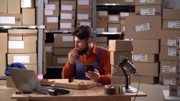 英俊的男性仓库工人一边坐在工作场所一边吃披萨 一边用他的智能手机为社交媒体服务 复制空间 — 图库视频影像