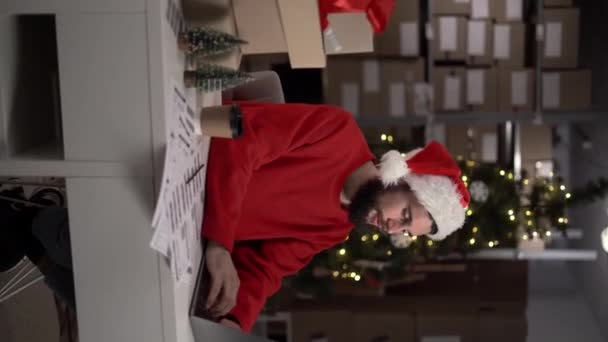 ノートパソコンのEコマースと小規模なビジネスコンセプトを使用して クリスマスのチェックアウトオンライン注文で倉庫で働く若い男 バーティカルビデオ — ストック動画