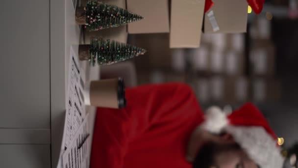 サンタの帽子の男は クリスマスイブの倉庫で働いています ラップトップを使用してビデオ通話は 配達中の顧客と通信します バーティカルビデオ — ストック動画