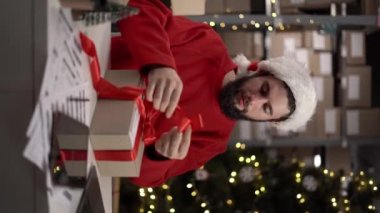 Noel Baba şapkalı bir adam teslimat için depoda Noel hediyeleri hazırlar ve kırmızı kurdele bağlar. Masada karton kutular var ve işadamı eşyalarını topluyor. Yeni yıl indirimi konsepti. Dikey video