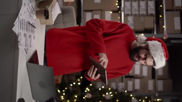 倉庫で働くサンタクロースの帽子と赤いセーターのビジネスオーナーを身に着けている白人のひげ付き男 クリスマスギフトショッピングコンセプト 配達のための箱 バーティカルビデオ — ストック動画
