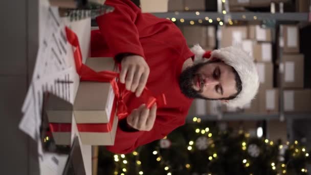 サンタの帽子の男は配達のための倉庫でクリスマスギフトを準備し 赤いリボンを結びます テーブルの上の段ボール箱とビジネスマンは梱包しています 新年のセールスコンセプト バーティカルビデオ — ストック動画
