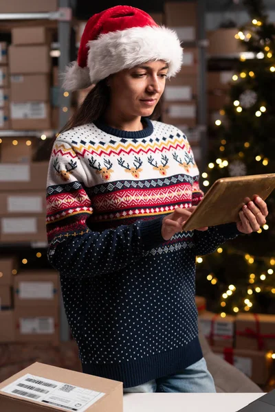 Female inventory manager holding digital tablet with Information about parcels. Christmas sale in e-shop. Working in stock of parcels with products ready for shipment. Copy space