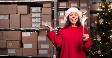 Elinde Mutlu Noeller havai fişekleri olan bir kadın atölye deposunda kameraya bakıp gülüyor. Hızlı Noel teslimatı konsepti. Pankart. Boşluğu kopyala