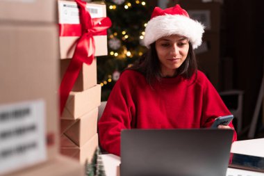 Teslimat işi. Bilgisayarı ve paketleri olan mutlu bir kadın depoda akıllı telefon uygulaması kullanarak Noel indirimi kutuları hazırlıyor. Boşluğu kopyala