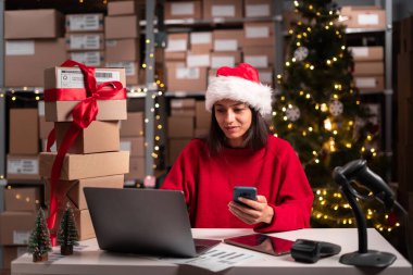 Noel 'de çalışan depo müdürü akıllı telefon ve dizüstü bilgisayar kullanıyor, internetten sipariş alıyor, Noel Baba şapkasındaki depo çalışanı mağazasından sipariş alıyor. Boşluğu kopyala