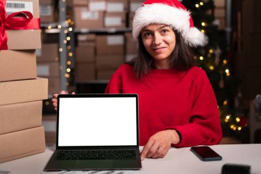 Depo çalışanı kadın Noel Baba şapkası takıyor. Depoda masasında beyaz boş ekran var. Kameraya bakıyor ve gülümsüyor. Boşluğu kopyala