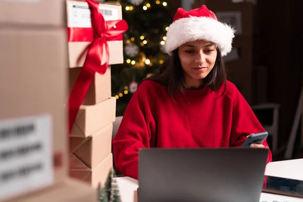 Delivery business. Happy woman with laptop and parcels using smartphone app in the warehouse preparing Christmas sale boxes. Copy space