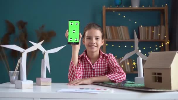 風力タービンと携帯電話を使用して自宅で代替の再生可能エネルギークリエイティブプロジェクトを行っている女子高生は カメラに緑色のスクリーン携帯電話を示しています コピースペース — ストック動画