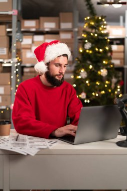Mutlu Noel Baba, Noel 'de atölye masasında dizüstü bilgisayar kullanarak şapka takıyor. İnternet sitesi, online alışveriş konsepti. Boşluğu kopyala