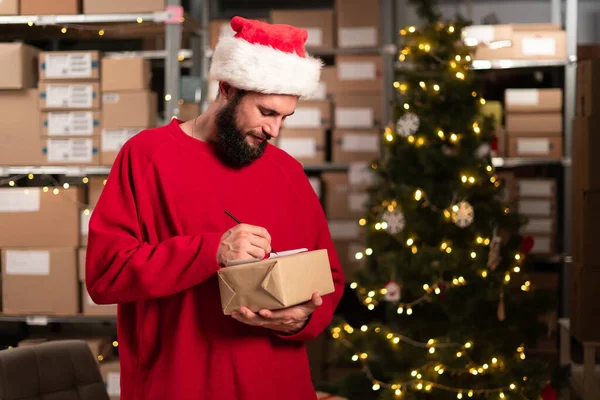 ハッピーサンタクロースパッキングプレゼントボックスのライティングアドレスは ワークショップの倉庫で郵送配達小包を準備します メリークリスマス配送コンセプト — ストック写真